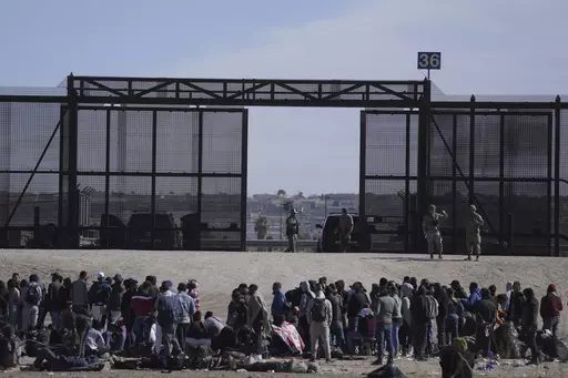Migrants who crossed the border from Mexico into the U.S. wait next to the U.S. border wall where U.S. Border Patrol agents stand guard, seen from Ciudad Juarez, Mexico, Thursday, March 30, 2023. The Biden administration will open migration centers in South and Central America for asylum seekers heading to the U.S.-Mexico border, in a bid to slow what’s expected to be a surge of migrants seeking to cross the border next month as pandemic-era immigration restrictions end, U.S. officials said Th