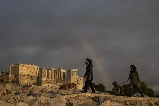 In this Wednesday, March 31, 2021, file photo, women wearing face masks walk with their dogs at Areopagus hill, in front of ancient Acropolis hill, as a rainbow is seen in the cloudy Athenian sky. Greece's culture ministry announced on Thursday, April 27, 2023, that pets will soon be allowed into more than 120 archaeological sites across the country, although not in some of the top tourist draws such as the Acropolis in Athens. (AP Photo/Petros Giannakouris, File)