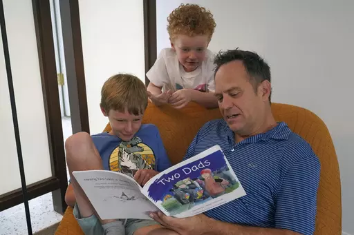 Wes Brown, right, reads to children Shawn Larimer-Brown, 7, left, and Charlie Larimer-Brown, 5, center, at their home Friday, Aug. 18, 2023, in Winter Park, Fla. Across the country, books and lessons that represent different families and identities to the youngest of learners are increasingly the target of the conservative pushback to efforts to promote diversity and inclusion. (AP Photo/John Raoux)