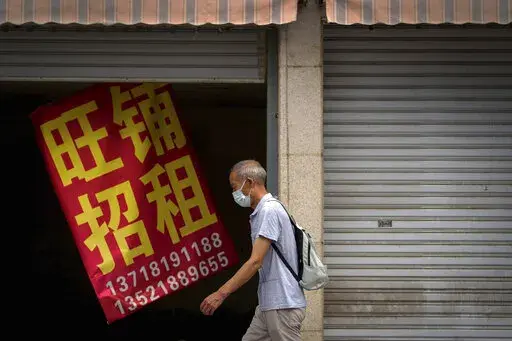 A man wearing a face mask walks by a banner that reads "Prosperous shop for rent" hangs on a vacant shop lot in Beijing, Wednesday, Aug. 17, 2022. Factories in China's southwest have shut down after reservoirs used to generate hydropower ran low in a worsening drought, adding to economic strains at a time when President Xi Jinping is trying to extend his position in power. (AP Photo/Andy Wong)