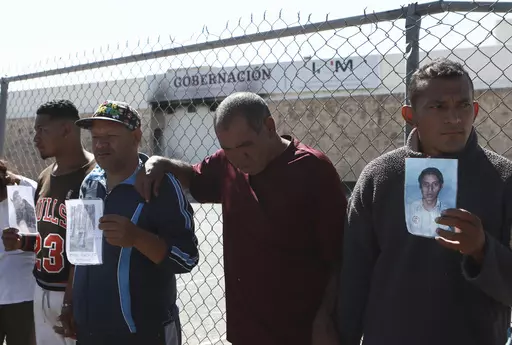Migrants, mostly from Venezuela, hold photos of those who died in a fire at a Mexican immigration detention center, behind, during a prayer vigil outside the center in Ciudad Juarez, Mexico, April, 27, 2023. Four months after a fire in a Mexican immigration detention center at the border killed 40 migrants, some survivors are living in limbo at a Mexico City hotel, recovering from their injuries and awaiting the prosecution of their captors. (AP Photo/Christian Chavez, File)