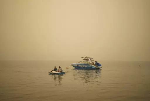 Smoke fills the air as Pat Manzuik and her husband Trevor use a paddleboat to get to shore after being given a boat ride by good samaritan Christy Dewalt, back right, back to their home they were evacuated from due to the Lower East Adams Lake wildfire, in Scotch Creek, Canada, Sunday, Aug. 20, 2023. (Darryl Dyck/The Canadian Press via AP)