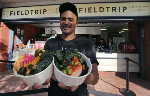 This Aug. 30, 2019 photo shows chef JJ Johnson posing with two signature rice bowls, one salmon and one vegetable, outside his Field Trip counter-service restaurant kiosk on the food court at the US Open tennis championships in New York. Johnson's new cookbook "The Simple Art of Rice: Recipes from Around the World for the Heart of Your Table," a book co-authored with Danica Novgorodoff, celebrates rice's versatility. (AP Photo/Charles Krupa, File)