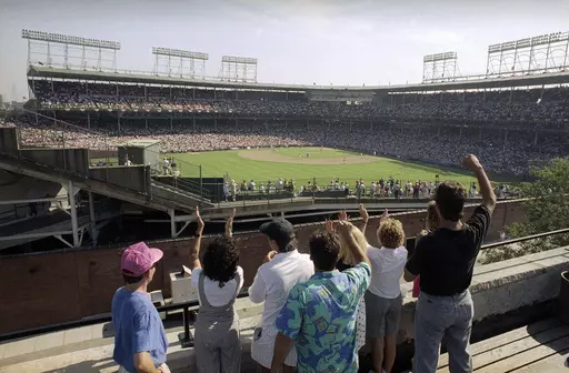 In this Monday, July 9, 1990, file photo, spectators watch an All-Star Game practice session from the roof of a building just outside Chicago's Wrigley Field. Booking hotels for baseball games during shoulder seasons like May, June or September can often be cheaper than the summer months. Additionally, save money by planning to attend weekday versus weekend games. (AP Photo/Seth Perlman, File)