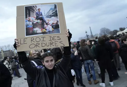 A woman holds a placard depicting French President Emmanuel Macron sitting on garbage cans that reads, "king of trash" during a protest in Paris, Friday, March 17, 2023. Protests against French President Emmanuel Macron's decision to force a bill raising the retirement age from 62 to 64 through parliament without a vote disrupted traffic, garbage collection and university campuses in Paris as opponents of the change maintained their resolve to get the government to back down. (AP Photo/Lewis Jol